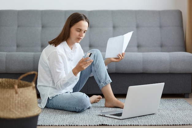 Imagem de jovem adulta séria vestindo camisa branca e jeans sentado no chão segurando documento em papel e celular na mão pagando contas de serviços públicos online