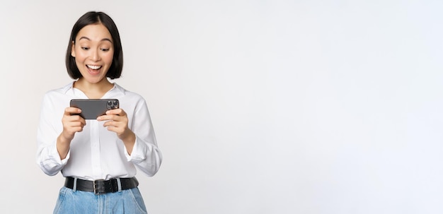 Imagem de jovem asiática assistindo no aplicativo de smartphone segurando o celular e olhando para a tela rindo e sorrindo em pé sobre fundo branco