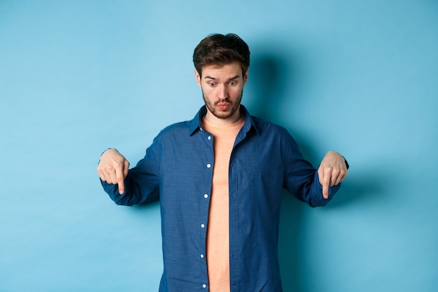 Imagem de jovem chocado em roupas casuais, olhando e apontando para algo estranho, de pé sobre um fundo azul.
