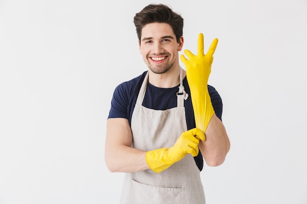 Imagem de jovem europeu usando luvas de borracha amarela para proteção das mãos sorrindo enquanto limpa uma casa isolada sobre o branco