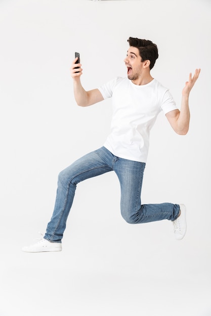 Imagem de jovem feliz animado posando isolado sobre uma parede branca, usando o telefone móvel pulando.