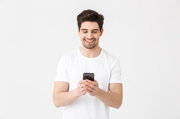 Imagem de jovem feliz animado posando isolado sobre uma parede branca, usando telefone celular.