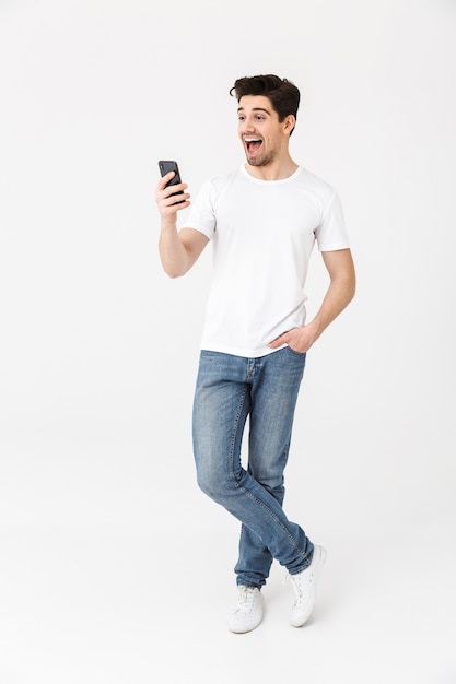 Imagem de jovem feliz animado posando isolado sobre uma parede branca, usando telefone celular.