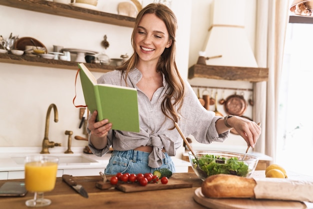 Imagem de jovem feliz sorridente alegre bonita dentro de casa na cozinha cozinhando segurando o caderno.