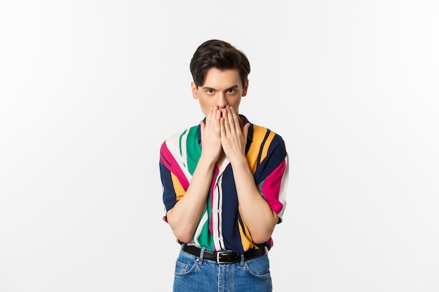 Imagem de jovem gay parecendo intrigado, segurando a boca com as mãos, fofocando, de pé sobre o branco