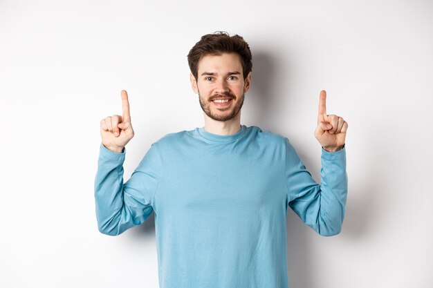 Imagem de jovem moderno com barba mostrando anúncio no espaço da cópia, apontando os dedos para cima e sorrindo, olha só gesto, fundo branco.