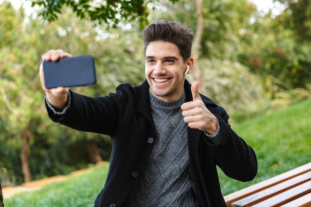 Imagem de jovem otimista em roupas casuais, caminhando ao ar livre no parque verde usando telefone celular, tire uma selfie com gesto de polegar para cima.