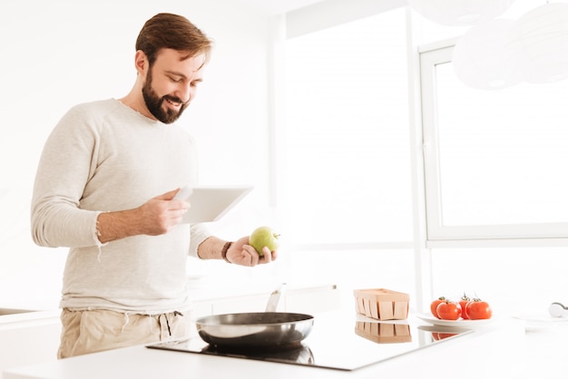 Imagem de jovem solteiro vestindo roupas casuais, cozinhando na cozinha, segurando a maçã e lendo a receita do computador tablet