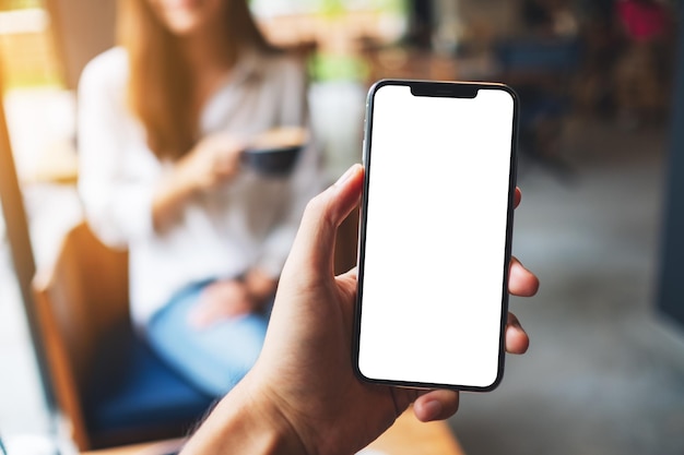 Imagem de maquete da mão de um homem segurando um telefone celular com uma tela em branco e uma mulher tomando café em um café