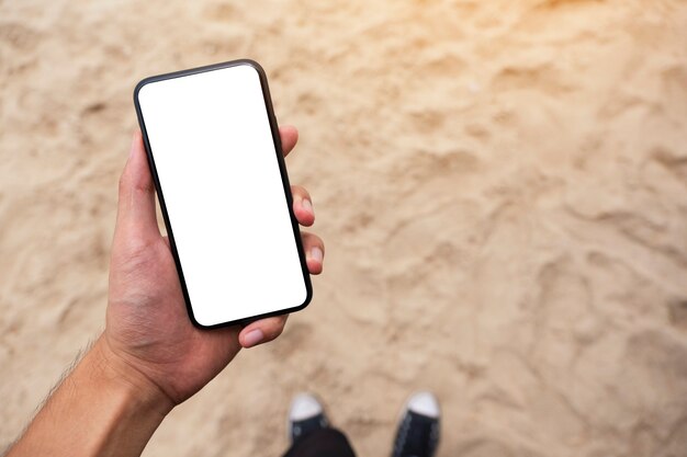 Imagem de maquete da mão de um homem segurando um telefone celular preto com uma tela em branco enquanto está de pé na praia