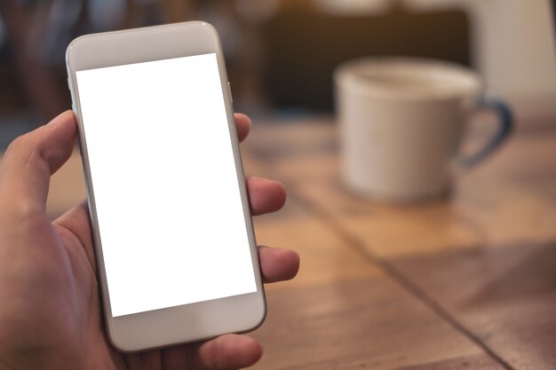 Imagem de maquete da mão segurando o celular branco com tela em branco com xícara de café na mesa de madeira no café