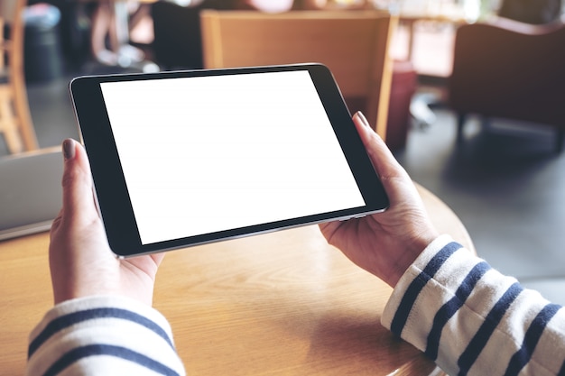 Imagem de maquete das mãos da mulher segurando preto tablet pc com tela de desktop branca em branco no café