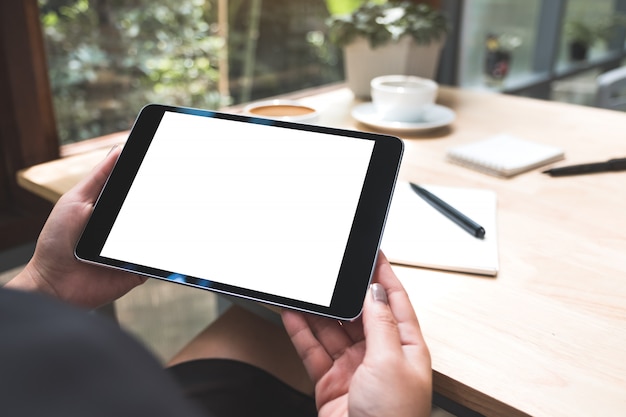 Imagem de maquete das mãos de uma mulher segurando preto tablet pc com tela em branco branca com notebook e xícara de café na mesa