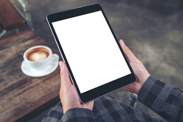 Imagem de maquete das mãos de uma mulher segurando um tablet pc preto com tela em branco e uma xícara de café na mesa de madeira no café