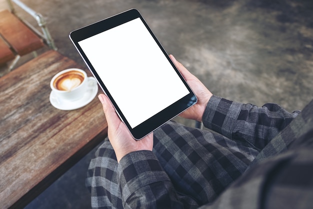 Imagem de maquete das mãos de uma mulher segurando um tablet pc preto com tela em branco e uma xícara de café