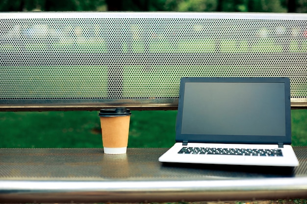 Imagem de maquete de laptop com tela preta em branco e xícara de café no banco de metal no parque natural ao ar livre. trabalho freelance.