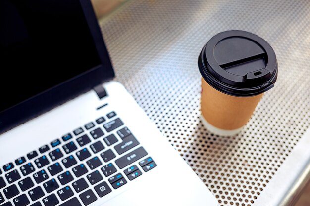 Imagem de maquete de laptop com tela preta em branco e xícara de café no banco de metal no parque natural ao ar livre. Trabalho freelance.