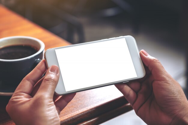 Imagem de maquete de mãos segurando e usando um celular branco com tela em branco horizontalmente para assistir com uma xícara de café na mesa de madeira