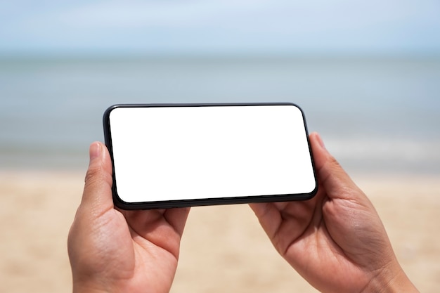 Imagem de maquete de mãos segurando um celular preto com uma tela em branco à beira-mar