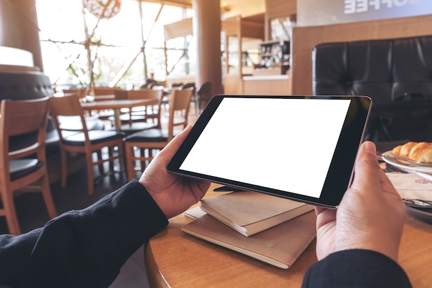 Imagem de maquete de mãos segurando um tablet pc preto com tela em branco com notebook e pão na mesa de madeira no café