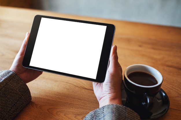 Imagem de maquete de mãos segurando um tablet pc preto com uma tela em branco e uma xícara de café na mesa de madeira