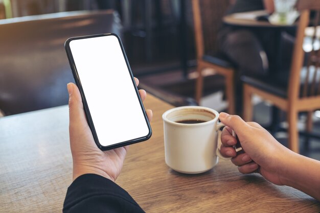 Imagem de maquete de mãos segurando um telefone celular branco com tela em branco enquanto bebe café em um café moderno