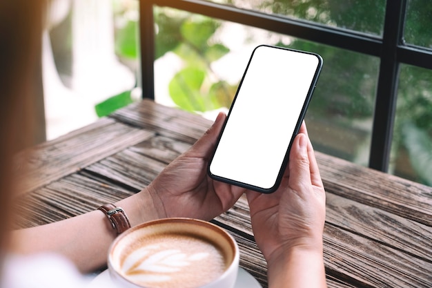 Imagem de maquete de mãos segurando um telefone celular preto com uma tela em branco e uma xícara de café em cima da mesa