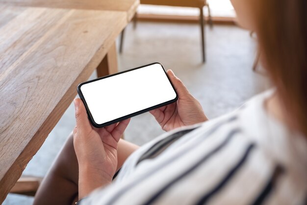 Imagem de maquete de mulher segurando um celular preto com uma tela em branco horizontalmente em um café