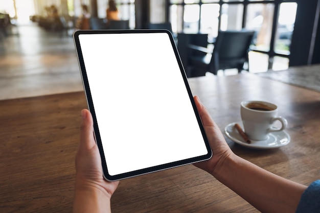 Imagem de maquete de uma mulher segurando o tablet digital com tela branca em branco do desktop com uma xícara de café na mesa de madeira