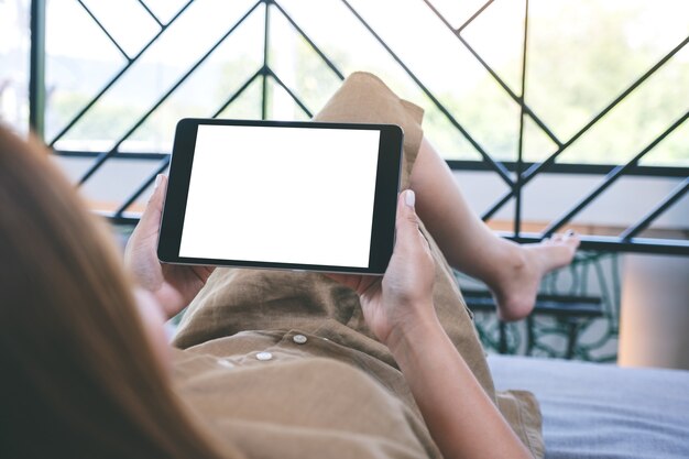 Imagem de maquete de uma mulher segurando um tablet pc preto com uma tela de desktop em branco enquanto está deitada na sala de estar sentindo-se relaxada