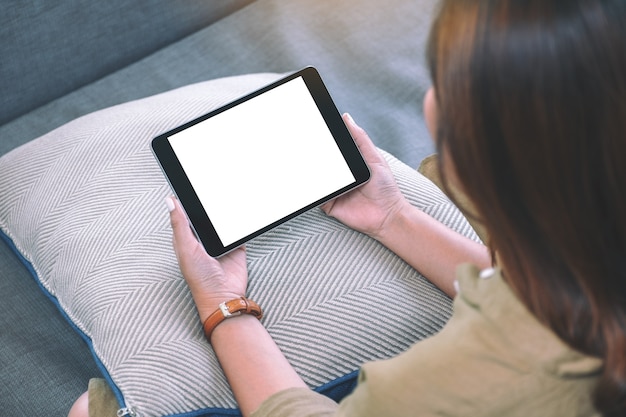 Imagem de maquete de uma mulher segurando um tablet pc preto com uma tela de desktop em branco enquanto está sentada na sala de estar sentindo-se relaxada
