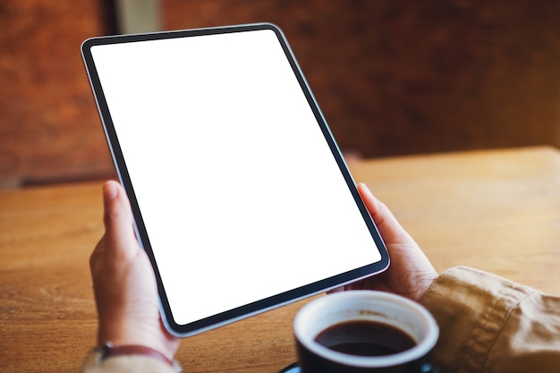 Imagem de maquete de uma mulher segurando um tablet pc preto com uma tela em branco e uma xícara de café na mesa de madeira