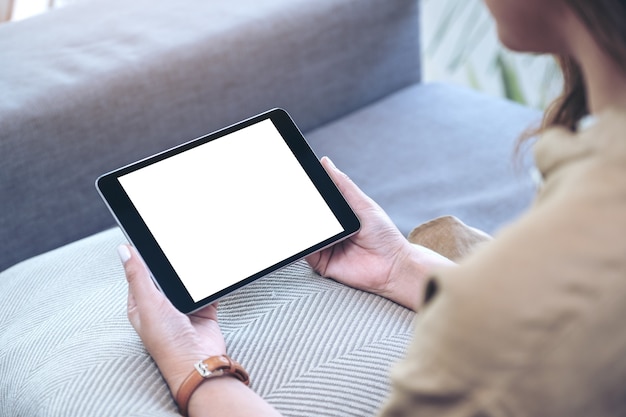 Imagem de maquete de uma mulher segurando um tablet pc preto com uma tela em branco na área de trabalho horizontalmente enquanto está sentada na sala de estar sentindo-se relaxada