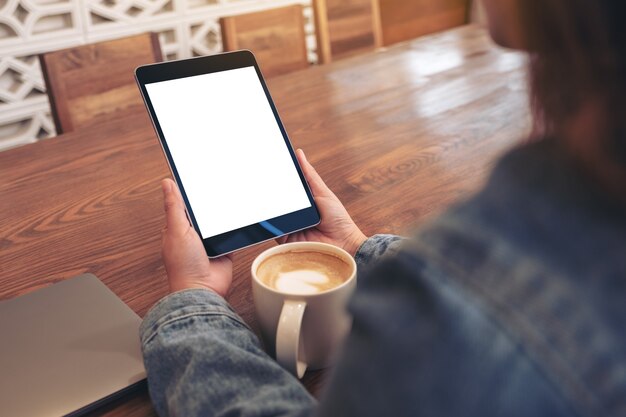 Imagem de maquete de uma mulher segurando um tablet pc preto com uma tela em branco, uma xícara de café e um laptop na mesa de madeira