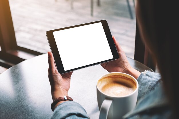 Imagem de maquete de uma mulher segurando um tablet preto com uma tela branca em branco enquanto bebe café