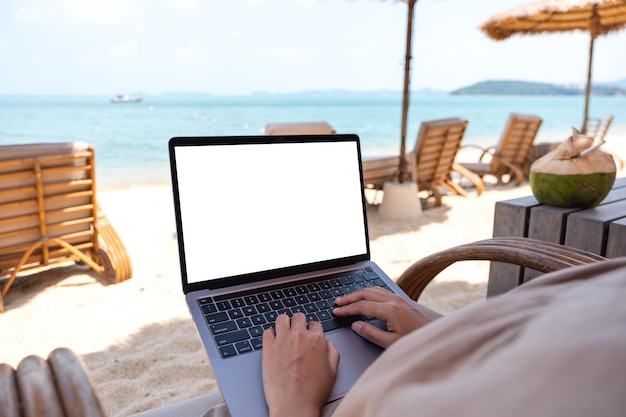 Imagem de maquete de uma mulher usando e digitando no computador portátil com tela em branco do desktop enquanto está sentado na praia