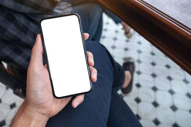 Imagem de maquete de vista superior da mão de um homem segurando um telefone celular branco com uma tela em branco na coxa em um café