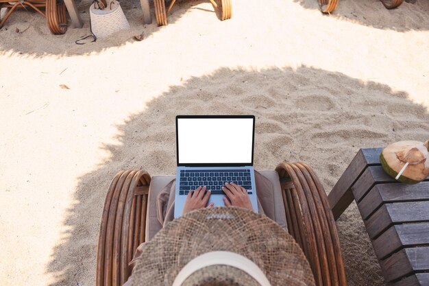 Imagem de maquete de vista superior de uma mulher usando e digitando no computador portátil com tela em branco do desktop enquanto está sentado na praia