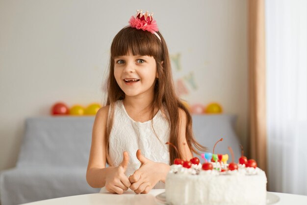 Imagem de menina bonitinha sentada à mesa com bolo de aniversário em casa contra um pano de fundo de balões olhando para a câmera com expressão feliz animada expressando felicidade