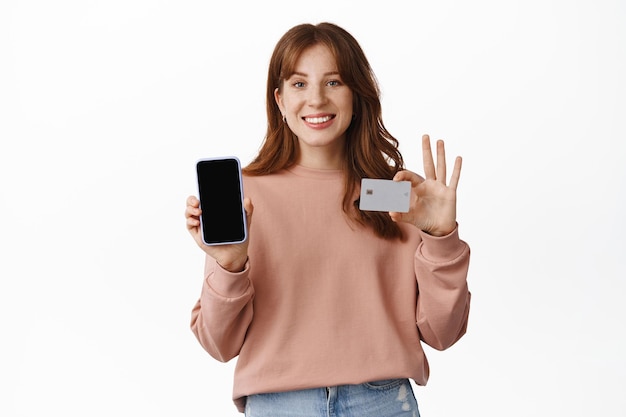 Imagem de menina ruiva sorridente mostrando a tela do celular e cartão de crédito, parecendo feliz, pedido de compras ou finanças, em pé contra um fundo branco