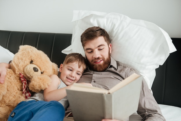 Imagem de menino encontra-se em casa, na cama, perto de um brinquedo com seu pai barbudo, lendo um livro de contos de fadas.