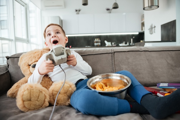 Imagem de menino no sofá com o ursinho de pelúcia em casa jogando no console enquanto come batatas fritas. segurando o joystick.