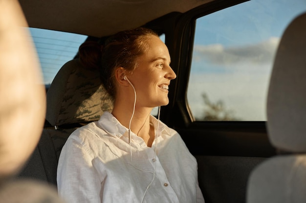 Imagem de mulher alegre alegre feliz vestindo camisa branca sentada no banco de trás do carro e ouvindo música em fones de ouvido olhando para longe na expressão positiva sorrindo
