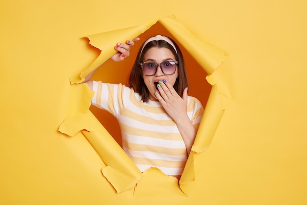 Imagem de mulher bonita caucasiana usando óculos de sol de faixa de cabelo e camiseta listrada em pé no buraco de papel amarelo sendo surpreendido e espantado cobrindo a boca com a palma da mão