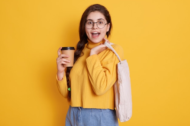 Imagem de mulher bonita e elegante segurando saco ecológico e levar café, senhora com expressão facial animada, vestindo roupas casuais e óculos.