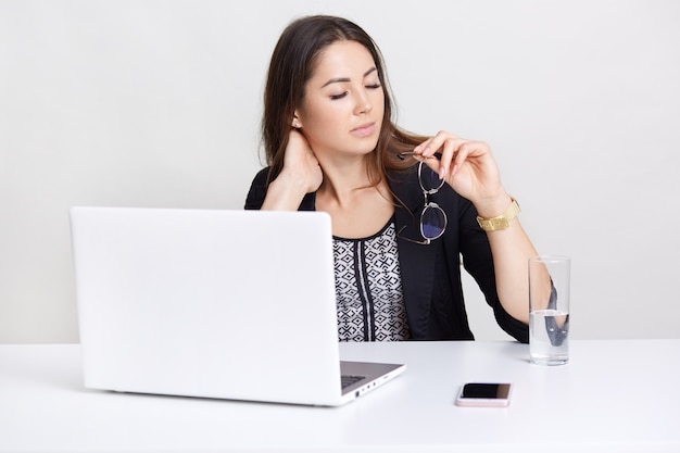 Imagem de mulher caucasiana cansada trabalha longas horas no computador portátil, mantém óculos na mão, sente dor no pescoço, rodeado de tecnologias modernas, senta-se na mesa branca, posa em ambiente interno