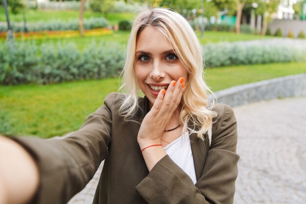 Imagem de mulher caucasiana de 20 anos vestindo uma jaqueta sorrindo e tirando uma foto de selfie enquanto caminha pelo parque