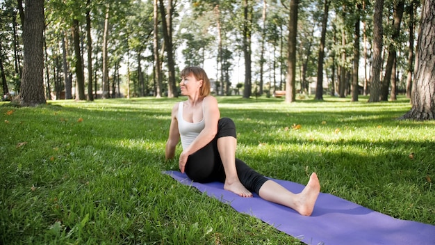 Imagem de mulher de meia idade sorridente com roupas de fitness, fazendo alongamento e exercícios de ioga. Mulher meditando e praticando esportes na esteira de ginástica na grama do parque
