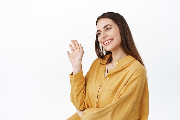 Foto imagem de mulher de pé meio virada acenando e olhando de lado cumprimentando alguém no espaço de cópia do lado esquerdo diga olá, olá, bem-vindo, pessoas em pé sobre fundo branco