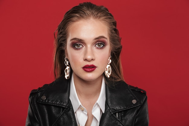 Imagem de mulher elegante e atraente de 20 anos com maquiagem brilhante, usando brincos e jaqueta de couro posando para a câmera sobre o vermelho
